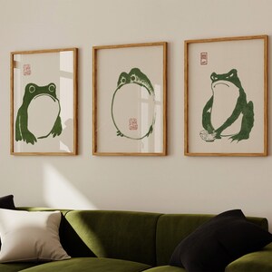 May include: Three framed prints of green frogs on a white background. The frogs are in different poses, one is sitting, one is standing, and one is in a circle. The frames are light brown wood.