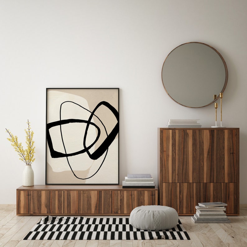 May include: A mid-century modern living room featuring a wooden cabinet, a round mirror, and a framed abstract print with black lines on a beige background. The cabinet has two doors and is crafted from wood with a natural finish. The mirror is round and has a gold frame. The print is framed in black and is hanging on the wall. A rug with a black and white chequered pattern is on the floor.