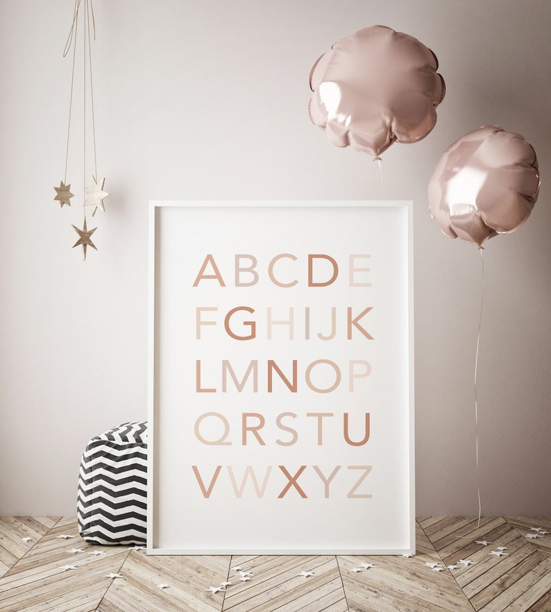 May include: A white framed poster with the alphabet in brown, pink, and beige letters on a white background. The poster is hanging on a white wall with two pink balloons and a wooden star mobile hanging above it.