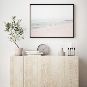 May include: A framed photograph of a beach scene with a light blue ocean and white sand. The photo is hanging on a white wall above a wooden cabinet with a potted plant, books, a clock, a vase with greenery, and a black candle holder.