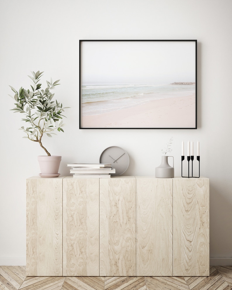 May include: A framed photograph of a beach scene with a light blue ocean and white sand. The photo is hanging on a white wall above a wooden cabinet with a potted plant, books, a clock, a vase with greenery, and a black candle holder.