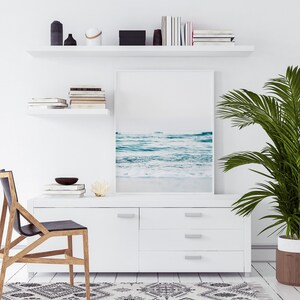 Peut inclure: Une commode blanche avec trois tiroirs et une photographie encadr&eacute;e d'une sc&egrave;ne de plage au-dessus. Un palmier en pot se trouve &agrave; droite de l'image. Une chaise en bois avec un si&egrave;ge noir est au premier plan.
