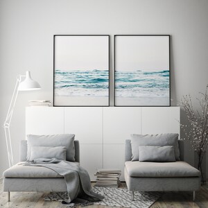 May include: Two framed photographs of a seascape with blue water and white foam waves. The photos are hanging on a white wall above a white cabinet with two gray chaise lounges in front of it.