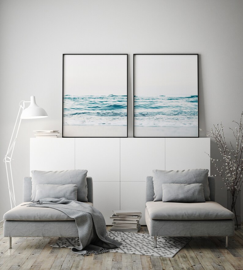 May include: Two framed photographs of a seascape with blue water and white foam waves. The photos are hanging on a white wall above a white cabinet with two gray chaise lounges in front of it.