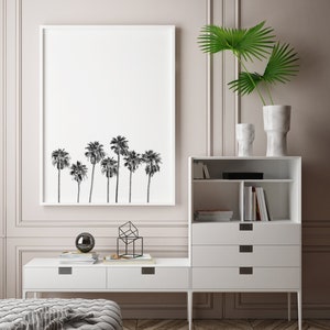 May include: A white framed print of a black and white silhouette of palm trees hangs on a light pink wall above a white media console with drawers and shelves. A geometric metal sculpture and a black orb sit on the console.