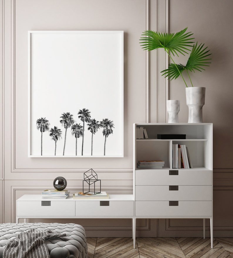May include: A white framed print of a black and white silhouette of palm trees hangs on a light pink wall above a white media console with drawers and shelves. A geometric metal sculpture and a black orb sit on the console.