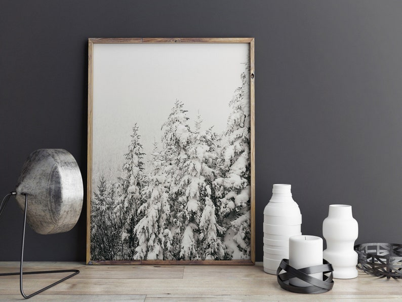 May include: A framed black and white photograph of snow-covered trees in a forest. The photo is hanging on a dark grey wall above a wooden table with white vases and a candle.