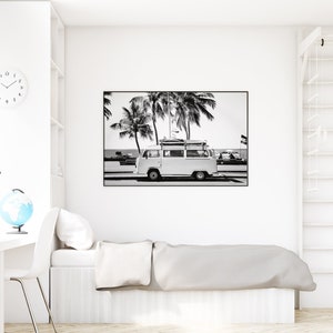 May include: Black and white photograph of a vintage van parked on a beach with palm trees in the background. The van has surfboards strapped to the roof.