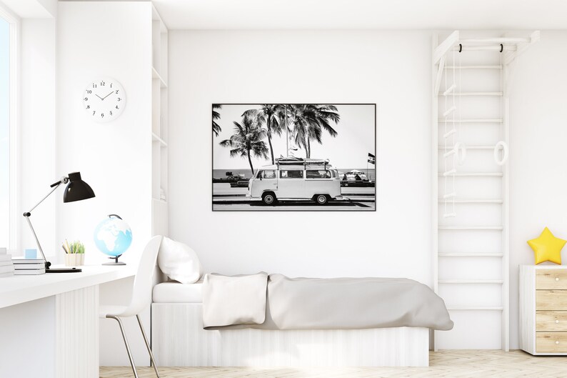 May include: Black and white photograph of a vintage van parked on a beach with palm trees in the background. The van has surfboards strapped to the roof.