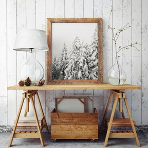 May include: A wooden table with two sawhorse style wooden stands. The table has a framed black and white photograph of a snowy forest, a lamp with a white shade, a clear glass vase with a branch of twigs, and a wooden crate with a canvas inside.