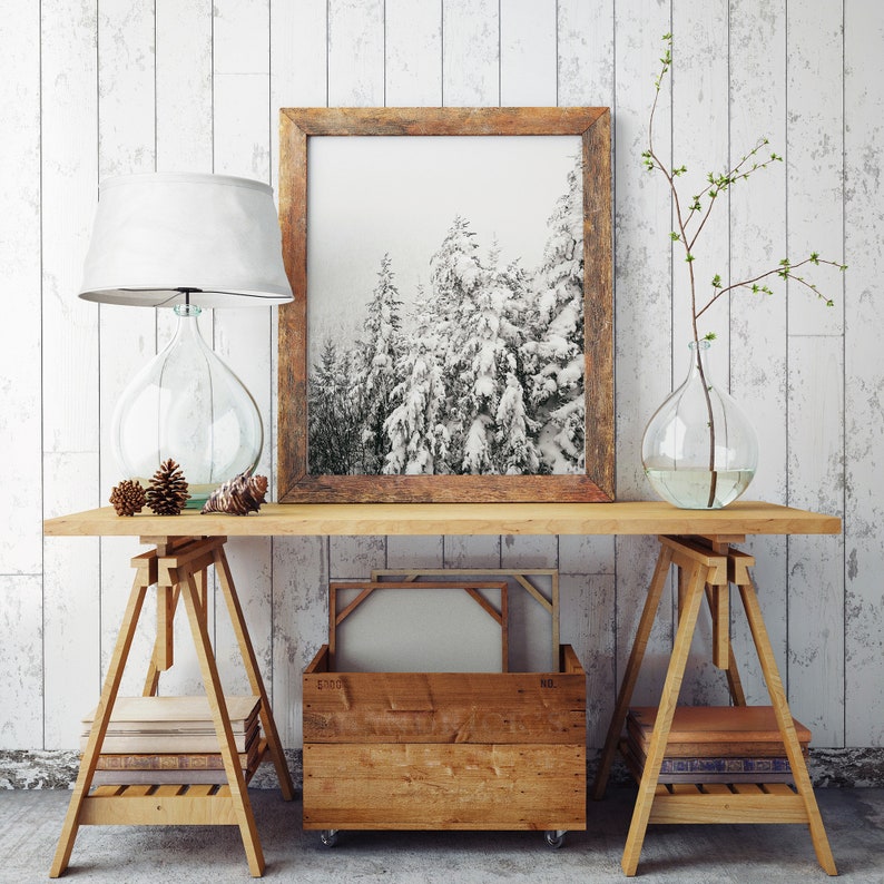 May include: A wooden table with two sawhorse style wooden stands. The table has a framed black and white photograph of a snowy forest, a lamp with a white shade, a clear glass vase with a branch of twigs, and a wooden crate with a canvas inside.
