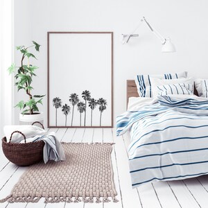 May include: A white bedroom with a bed with blue and white striped bedding. A framed print of palm trees hangs on the wall above the bed. A potted plant sits on the floor next to the bed.
