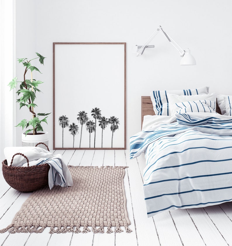 May include: A white bedroom with a bed with blue and white striped bedding. A framed print of palm trees hangs on the wall above the bed. A potted plant sits on the floor next to the bed.