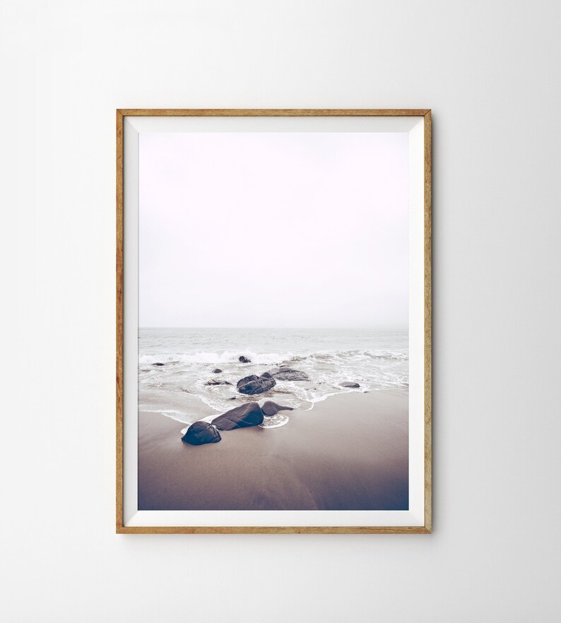 May include: A framed photograph of a beach scene with dark rocks and white waves crashing on the shore. The sky is a light gray and the sand is a light brown.