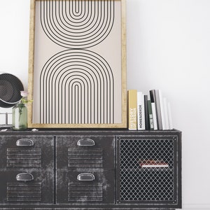 May include: A black and white geometric print in a wooden frame hangs above a black metal cabinet with four drawers and a wire mesh door. The cabinet is sitting on a white wood floor. There are several books stacked on top of the cabinet, including one titled "History of Design".
