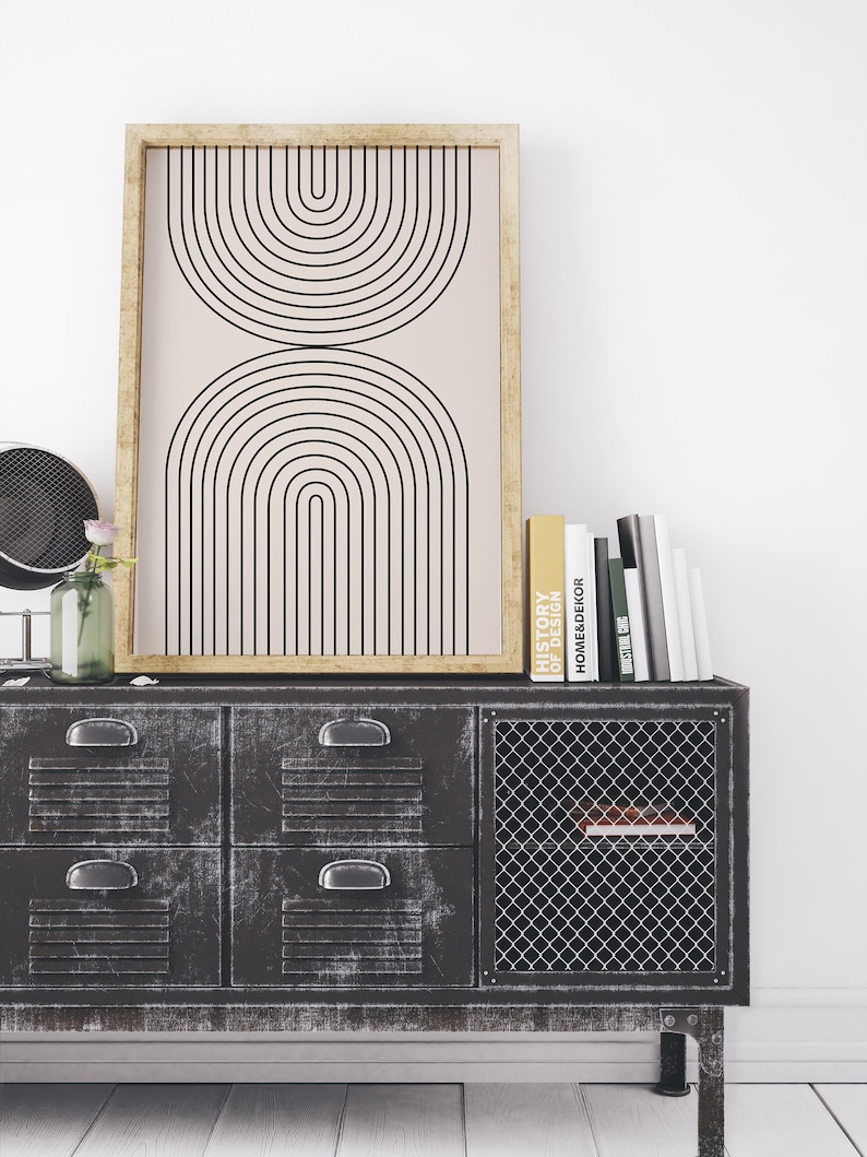 May include: A black and white geometric print in a wooden frame hangs above a black metal cabinet with four drawers and a wire mesh door. The cabinet is sitting on a white wood floor. There are several books stacked on top of the cabinet, including one titled "History of Design".