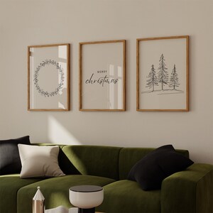 May include: Three framed prints with a minimalist Christmas theme. The left print features a wreath of leaves and berries. The center print says "Merry Christmas" in a simple script font. The right print shows three stylized pine trees.