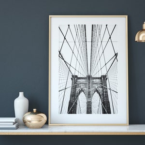 Pu&ograve; includere: Una fotografia in bianco e nero incorniciata del ponte di Brooklyn. La stampa &egrave; esposta su un tavolo con piano in marmo bianco e cornice dorata. Gli elementi decorativi includono un vaso bianco, un oggetto sferico dorato e libri impilati.