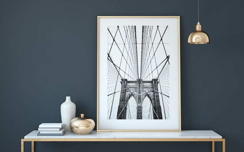 Pu&ograve; includere: Una fotografia in bianco e nero incorniciata del ponte di Brooklyn. La stampa &egrave; esposta su un tavolo con piano in marmo bianco e cornice dorata. Gli elementi decorativi includono un vaso bianco, un oggetto sferico dorato e libri impilati.