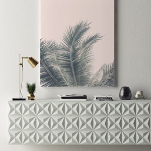 May include: A white geometric console table with a palm tree print framed artwork hanging above it. The artwork has a pink background and the palm tree leaves are green. There are two black books and a black and white book on top of the console table. There is a gold lamp with a black base and a small green plant in a gold pot on the left side of the console table. There is a black vase with a black and white pattern on the right side of the console table. There is a small white decorative object on the right side of the console table.