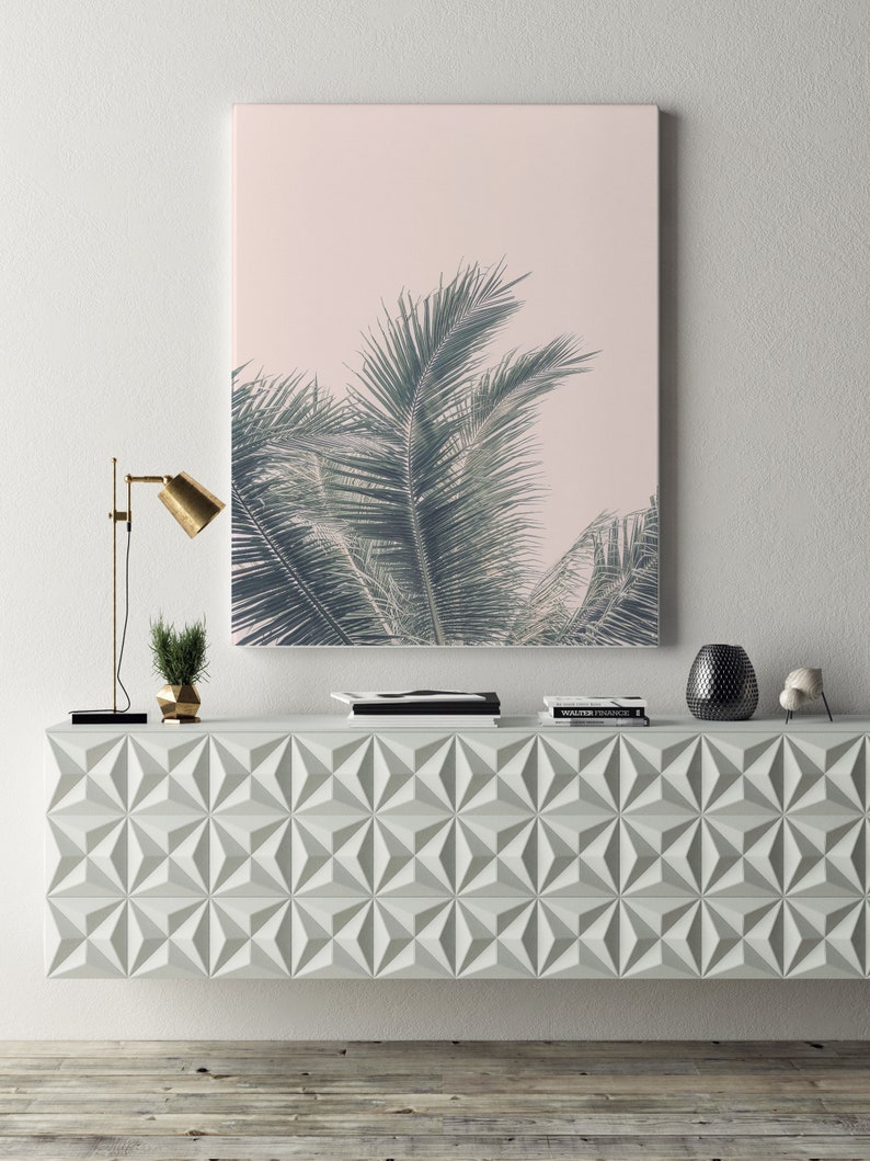 May include: A white geometric console table with a palm tree print framed artwork hanging above it. The artwork has a pink background and the palm tree leaves are green. There are two black books and a black and white book on top of the console table. There is a gold lamp with a black base and a small green plant in a gold pot on the left side of the console table. There is a black vase with a black and white pattern on the right side of the console table. There is a small white decorative object on the right side of the console table.