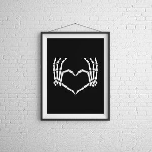 May include: Black and white print of two skeletal hands forming a heart shape. The print is framed in a black frame and hanging on a white brick wall.