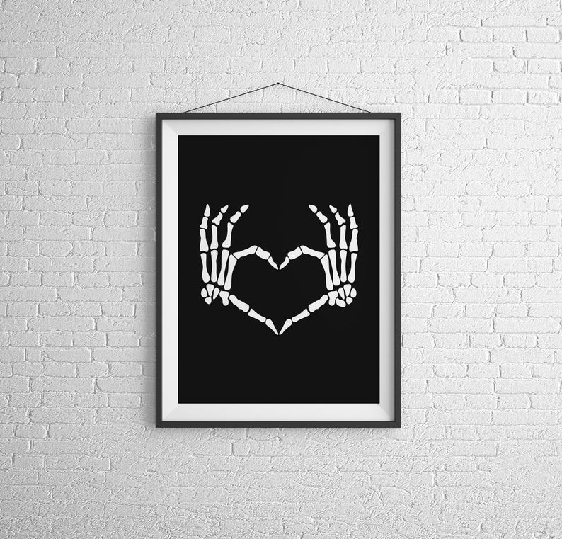 May include: Black and white print of two skeletal hands forming a heart shape. The print is framed in a black frame and hanging on a white brick wall.