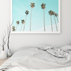 Puede incluir: Una impresi&oacute;n enmarcada de una fila de palmeras contra un cielo azul claro. La impresi&oacute;n est&aacute; colgada sobre una cama blanca con un edred&oacute;n gris.