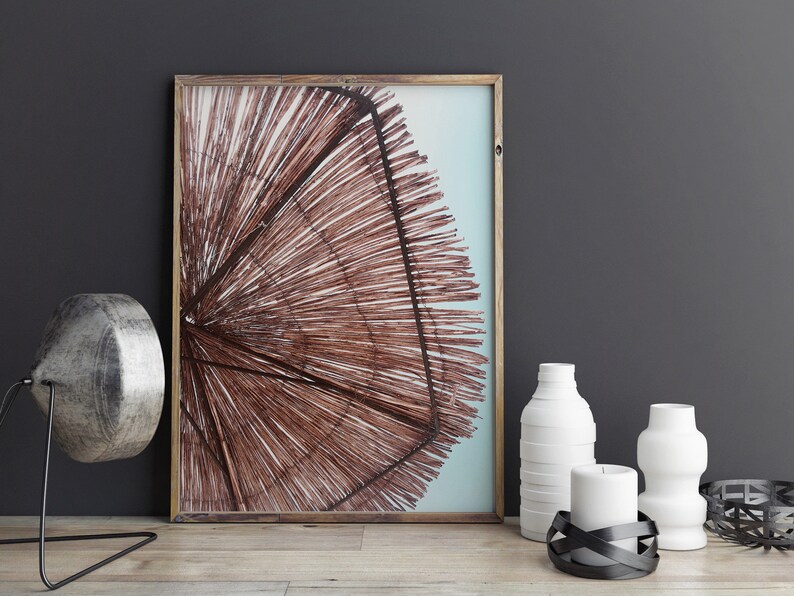 May include: A framed print of a brown woven beach umbrella against a blue sky. The print is on a table with a silver lamp, two white vases, a white candle, and a black metal candle holder.