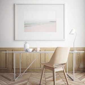 May include: A white framed picture of a beach scene hangs above a white table with a light wood top. A white ceramic pineapple and two bowls sit on the table. A beige chair sits at the table. A white lamp with a white shade sits on the table.