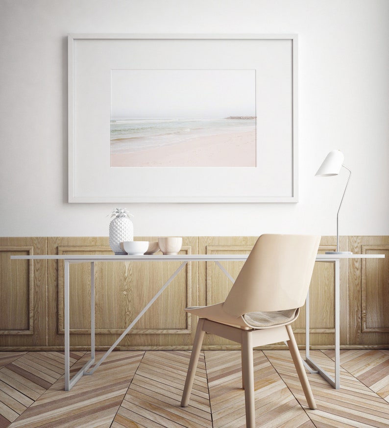May include: A white framed picture of a beach scene hangs above a white table with a light wood top. A white ceramic pineapple and two bowls sit on the table. A beige chair sits at the table. A white lamp with a white shade sits on the table.