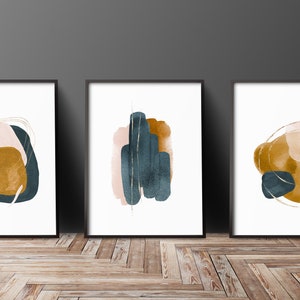 May include: Set of three abstract art prints featuring watercolour shapes in shades of blue, pink, and gold. The prints are framed in black frames and are displayed on a wooden floor.