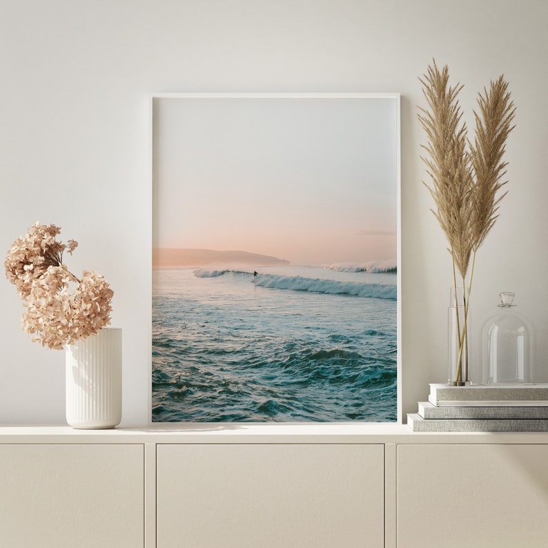 May include: A framed photograph of a seascape with a surfer riding a wave in the distance. The water is a light blue and green color, and the sky is a soft pink and orange.