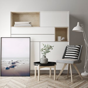 May include: A white shelf unit with a black framed photograph of a beach scene. The shelf unit has a wooden top and a small wooden shelf in the bottom right corner. A gray chair with a black and white striped cushion sits in front of the shelf unit. A small wooden table with a black top sits in front of the chair. A vase with green leaves sits on the table.