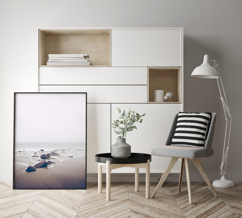 May include: A white shelf unit with a black framed photograph of a beach scene. The shelf unit has a wooden top and a small wooden shelf in the bottom right corner. A gray chair with a black and white striped cushion sits in front of the shelf unit. A small wooden table with a black top sits in front of the chair. A vase with green leaves sits on the table.