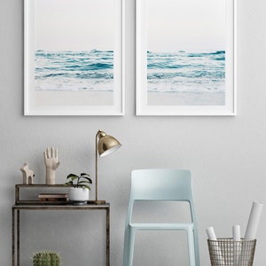 May include: Two framed photographs of ocean waves crashing on a sandy beach. The photos are hung on a white wall above a light blue chair, a side table with a lamp, and a potted cactus.