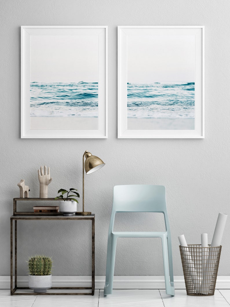 May include: Two framed photographs of ocean waves crashing on a sandy beach. The photos are hung on a white wall above a light blue chair, a side table with a lamp, and a potted cactus.