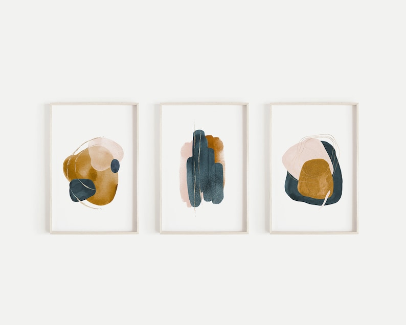 May include: Set of three abstract watercolour prints featuring muted tones of pink, gold, and blue. The prints are framed in a simple white frame.