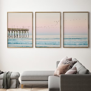 May include: A set of three framed prints depicting a seascape with a wooden pier extending into the ocean. The sky is a soft pink and blue, and there are birds flying overhead. The ocean is a light blue and turquoise colour, with white waves crashing on the shore.