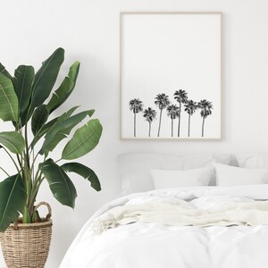 May include: A black and white print of palm trees in a simple wooden frame hanging on a white wall above a white bed with a white blanket and a green plant in a wicker basket.
