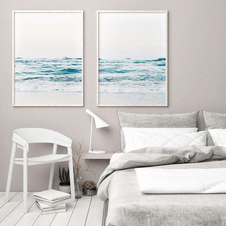 May include: Two framed prints of a seascape with blue water and white foam waves. The prints are hanging on a light gray wall above a white bed with gray bedding and white pillows.