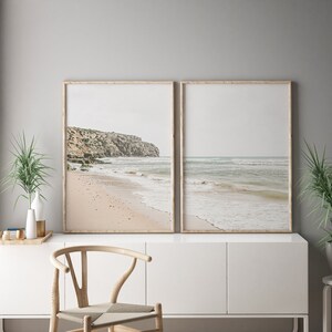 May include: Two framed beach prints. One print shows a sandy beach with a rocky cliffside. The other print shows the ocean with gentle waves. The frames are light brown, and the overall color palette is neutral with beige, gray, and blue tones.