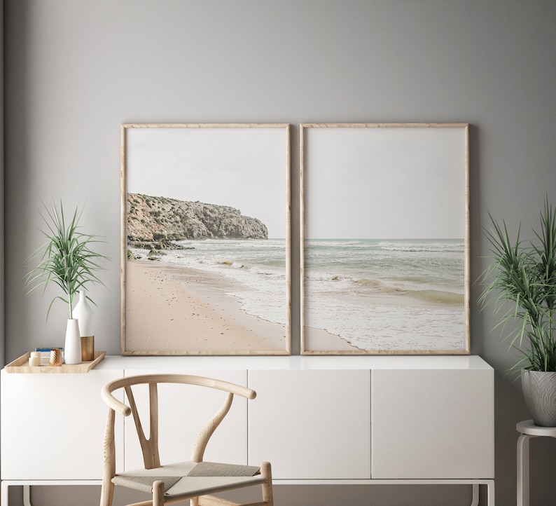 May include: Two framed beach prints. One print shows a sandy beach with a rocky cliffside. The other print shows the ocean with gentle waves. The frames are light brown, and the overall color palette is neutral with beige, gray, and blue tones.