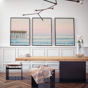 May include: Three framed photographs of a beach scene with a pier and birds flying overhead. The photos are hung on a white wall above a wooden table with a black metal base. There are two black metal stools with cushions on the floor in front of the table.
