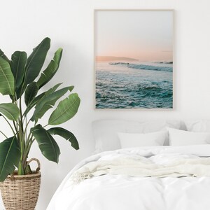 May include: A framed photograph of a seascape with a surfer riding a wave in the distance. The sky is a soft pink and orange, and the water is a turquoise blue.