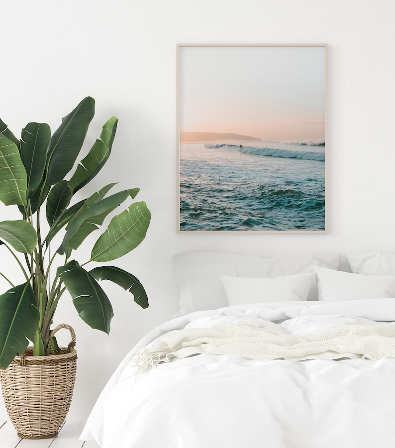 May include: A framed photograph of a seascape with a surfer riding a wave in the distance. The sky is a soft pink and orange, and the water is a turquoise blue.