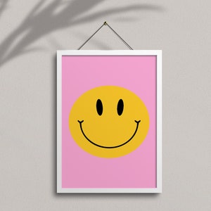 May include: A white framed print with a pink background and a yellow smiley face with black eyes and a black outline.