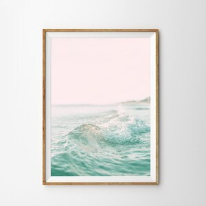May include: A framed photograph of a wave crashing on a beach. The water is a light blue-green color and the sky is a soft pink.