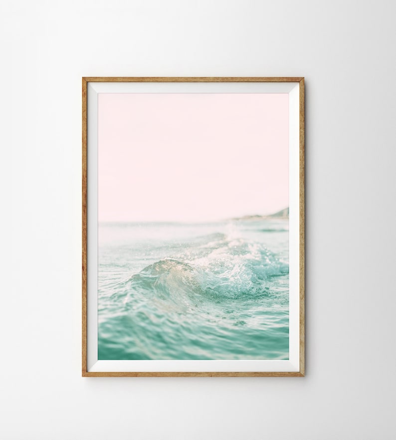 May include: A framed photograph of a wave crashing on a beach. The water is a light blue-green color and the sky is a soft pink.