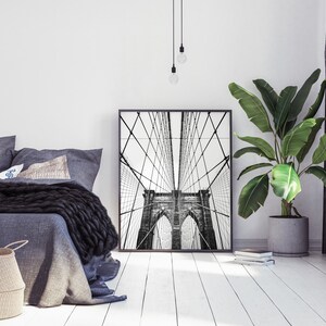 Pu&ograve; includere: Stampa incorniciata in bianco e nero del ponte di Brooklyn. L'opera d'arte &egrave; esposta in una camera da letto moderna con biancheria da letto grigia, un cesto di vimini e una pianta in vaso. L'ambiente &egrave; minimalista.